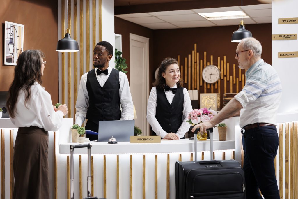 Two guests, elderly couple, arrive at hotel reception for check-in. Receptionists assists senior guests with booking and paperwork, creating welcoming atmosphere. Hotel lobby is lively with tourists.